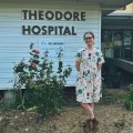 A woman in a dress and boots stands in front of a building which is labelled Theodore Hospital